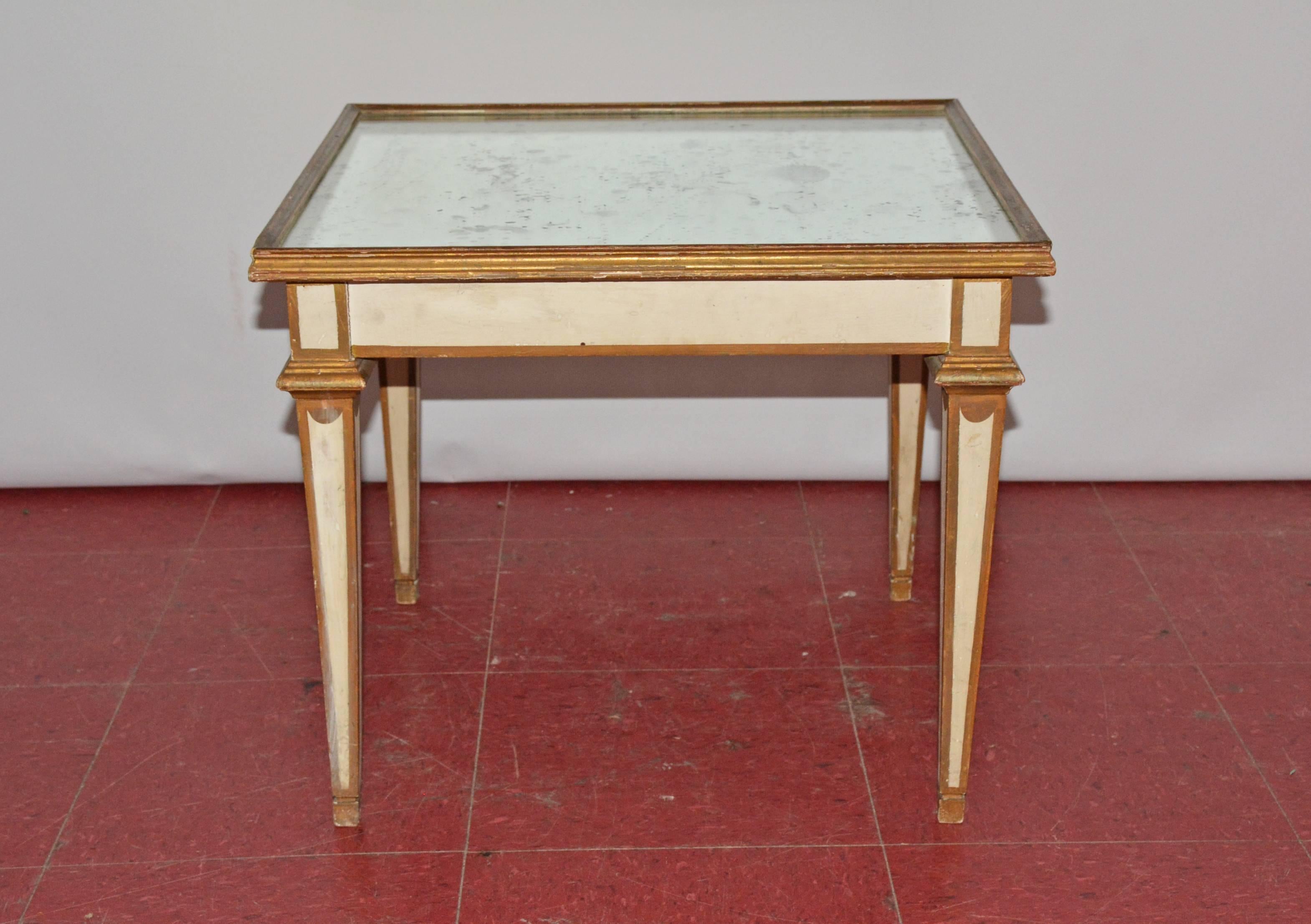 Painted white with gilt trim, the neoclassical coffee or end table features a mirrored top and tapered legs.