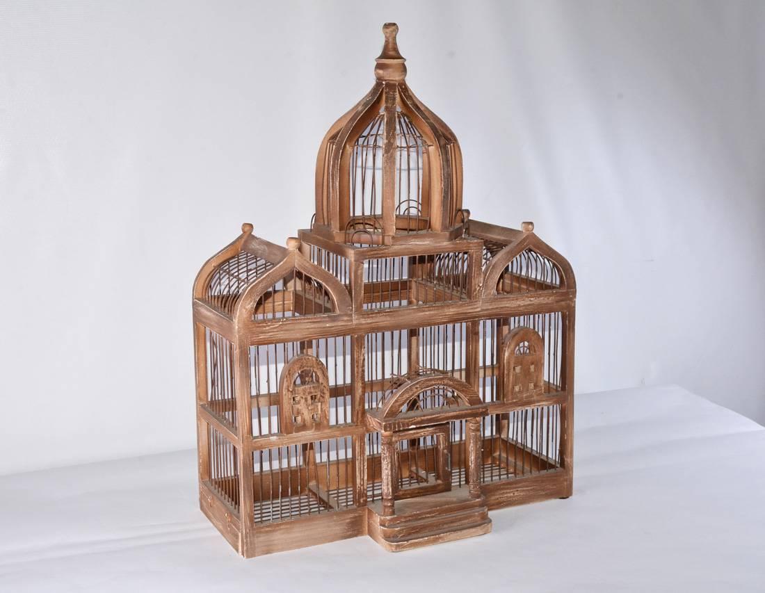The antique architectural bird cage has a pair of peaked roofs and tall centre dome under which is a pedimented portico leading to a swinging door. The frame is wood painted brown and is interspersed with wire caging. To the left at the bottom is a