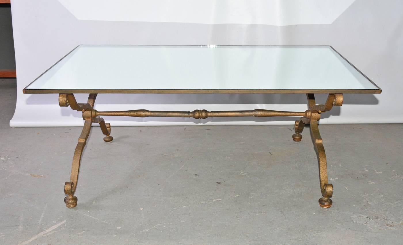 The glamorous rectangular Gilbert Poillerat style, circa 1940s vintage coffee or cocktail table has Rococo gilt wrought iron criss-cross braces for legs attached to a stretcher. The top is a mirror set within a gilt wrought iron frame.  Think 18th