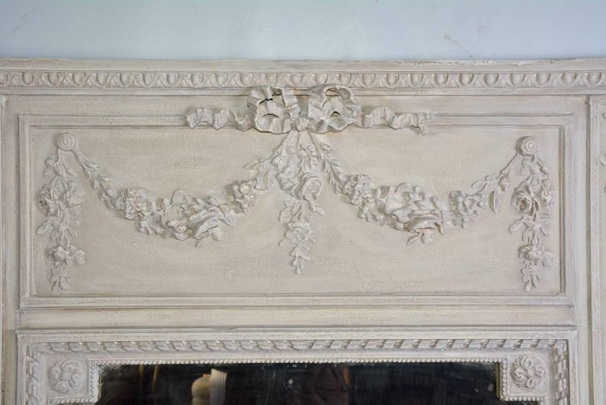 The antique French trumeau mirror is in the Louis XVI neoclassical style. Across the top is an egg-and-dart cornice and a floral garland tied with a rippling bow. The mirror glass is framed by a beaded border and ribbon border. The frame is painted