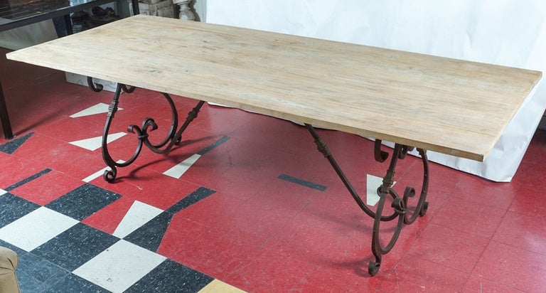 The Rococo or Baroque style table base shown with plank teak tabletop has a trestle base featuring two lyre-shaped legs adorned with a weathered rust color finish, connected to the underside of the top with a curved stretcher. The delicate volutes,