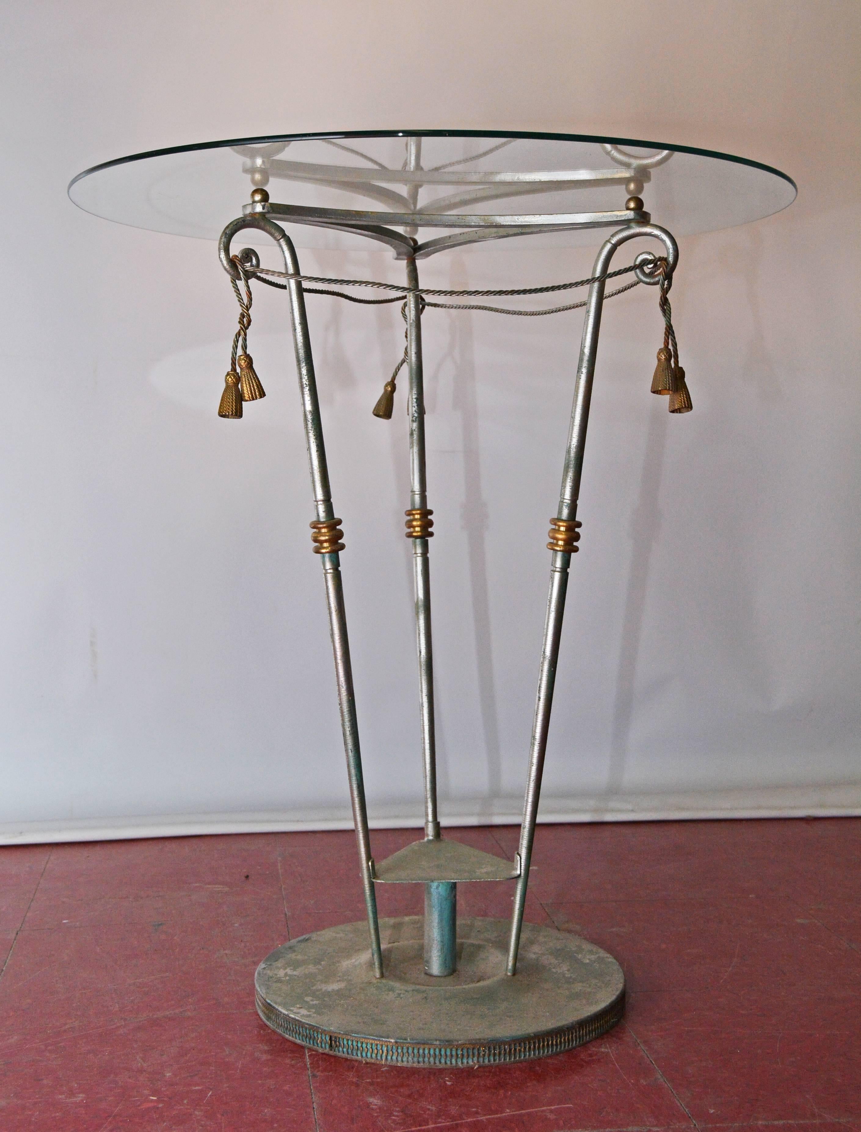 This pedestal center table has a round glass top that sits on an iron base which is both painted and gilded. The three angled legs are connected at the top by iron tasseled cords. Can be use as a lamp table or a plant stand for flower arrangement or