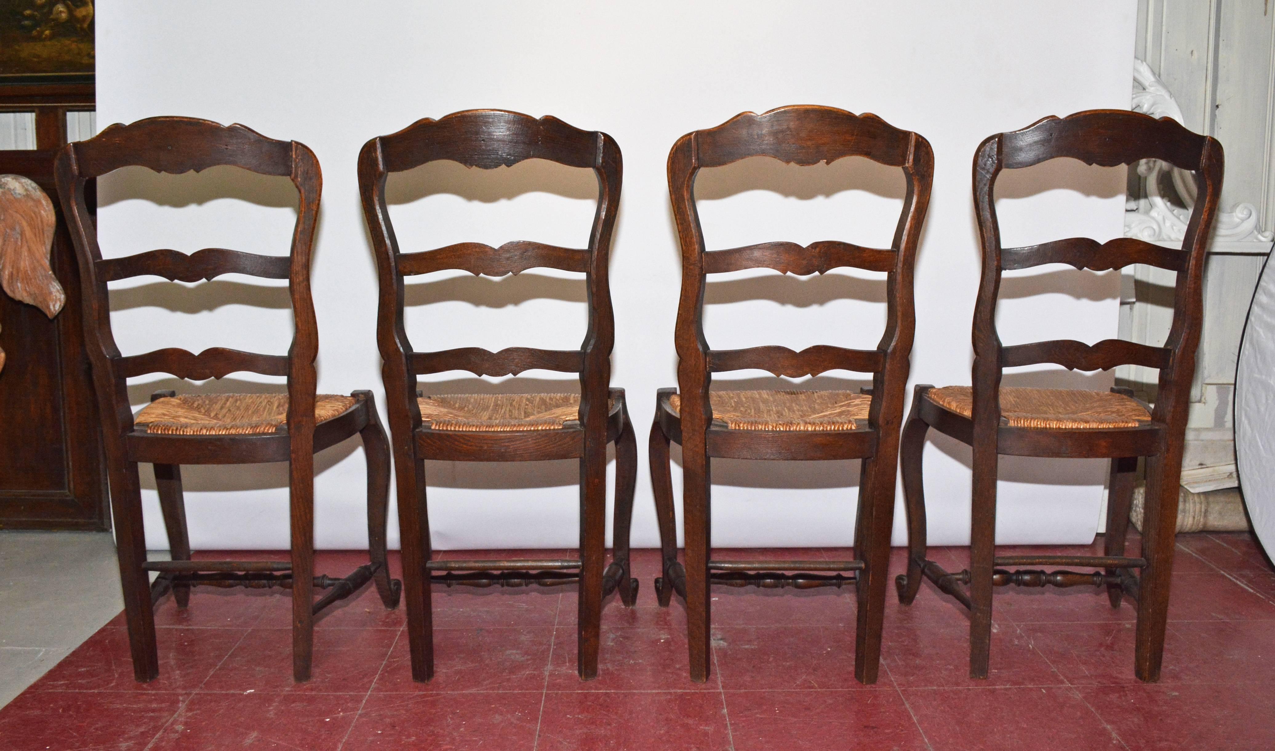 Set of four French Louis XV Provincial dining chairs have hand-carved shells on the crown of the ladder backs and on the seat aprons. There are cabriole legs and turned stretchers and rush seats.  Beautiful patina on antique oak wood.