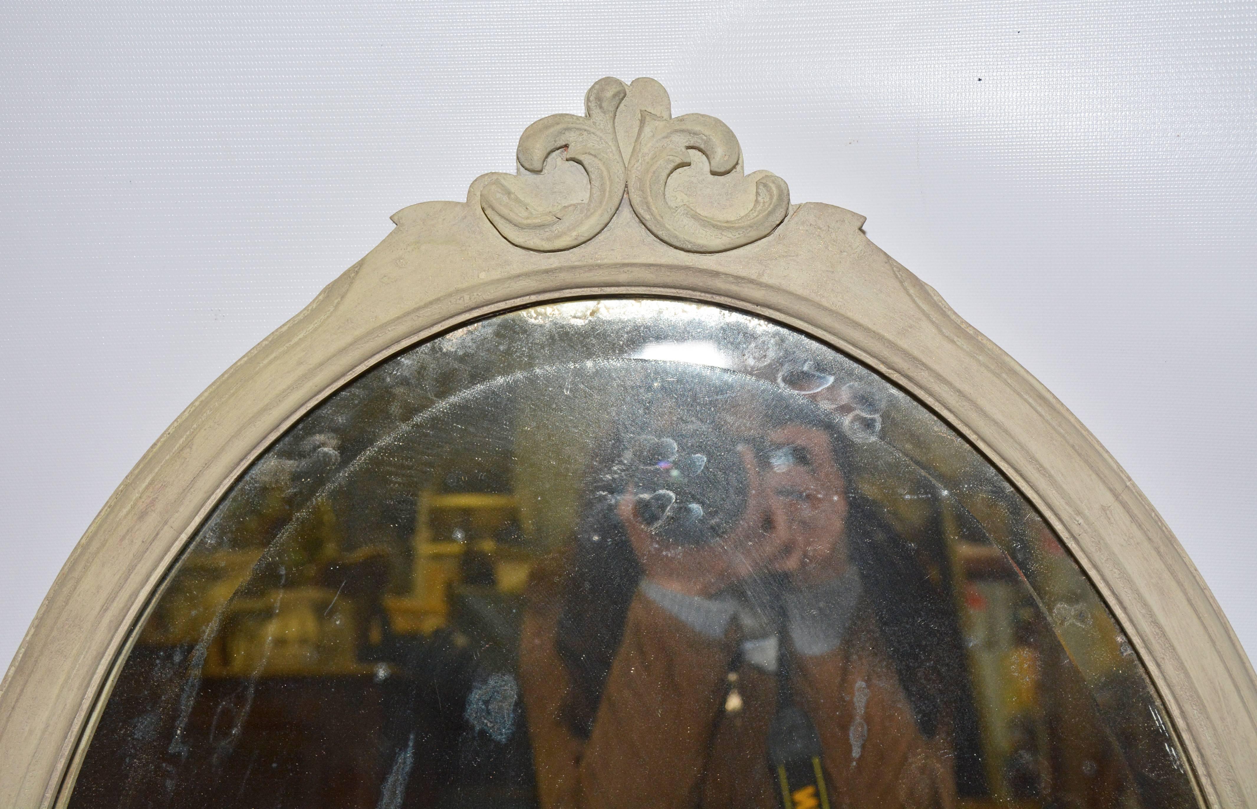 A fine oval frame mirror, painted light grey, is decorated at the top with two back-to-back scrolls. The silvered mirror is bevelled and the backing is wood and wired for hanging.