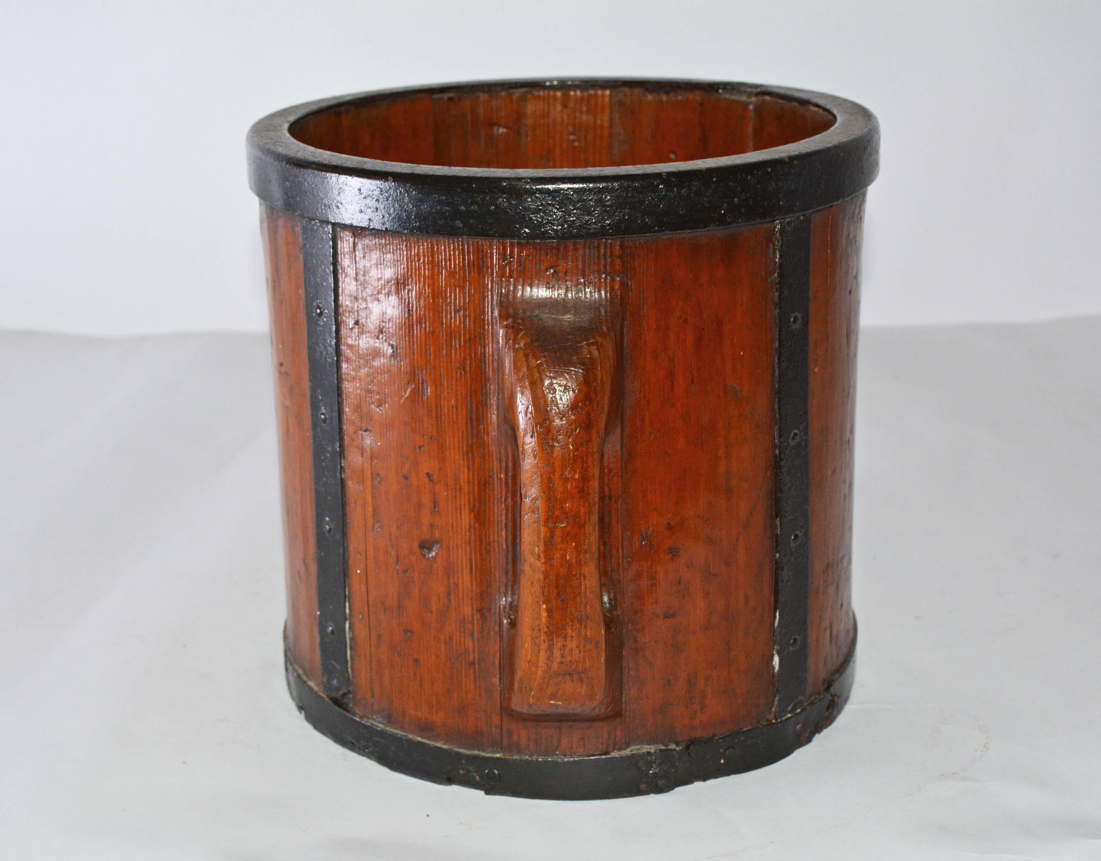 The antique Chinese wood bucket has two handles and iron straps on the sides and bottom. The surfaces are lacquered. Used originally for measuring rice or grain.