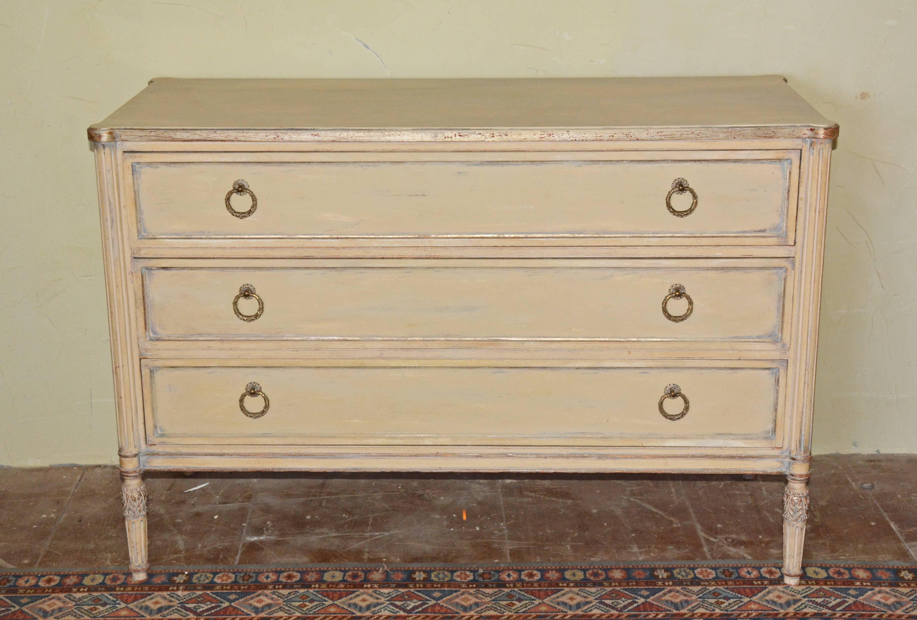 The Louis XVI-style, three-drawer chest or dresser is cream colored trimmed with silver gilt edging the top and framing the front of each drawer. Fluted legs and rounded fluted pilasters decorate each of the four corners. A pair of brass bay berry