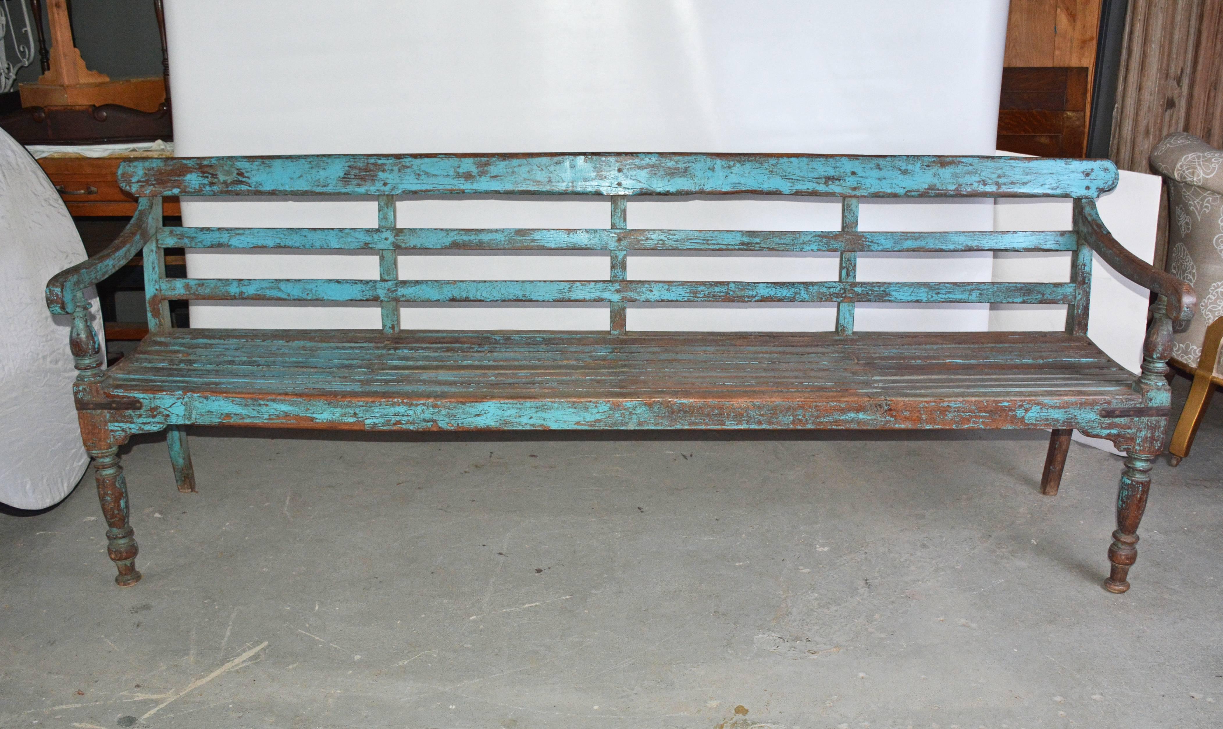 The antique blue-painted English Colonial style wood bench has slatted back and concave seat. Metal L-shaped straps secure all four corners. The front legs and arm supports are turned.
Measures: Arm height: 28".