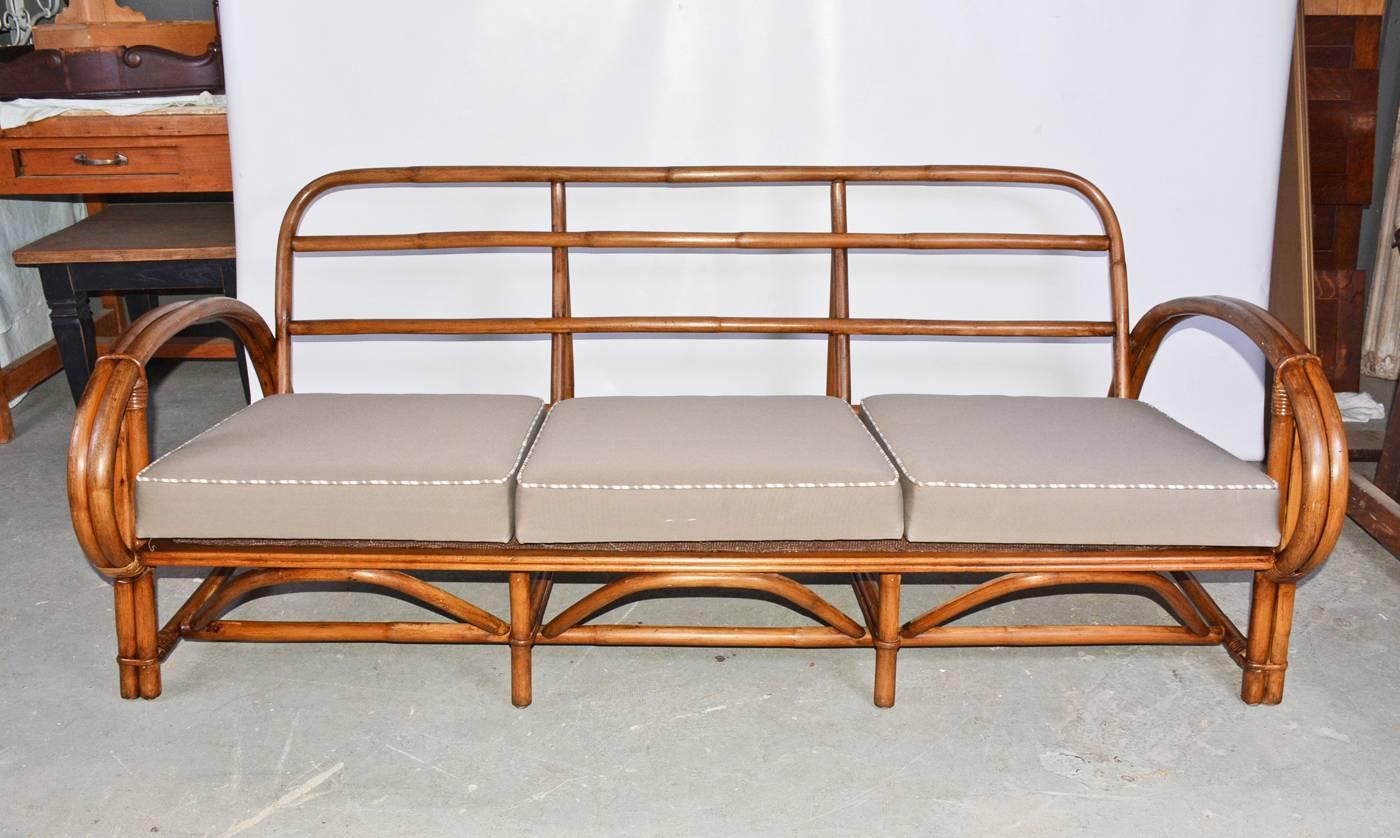 The vintage three-seat porch wicker rattan sofa with arms is made of thick brown rattan and sturdy construction with stretchers on all four sides and arched braces on the front. Each seat section has zig-zag iron-rod bracing to hold the upholstered