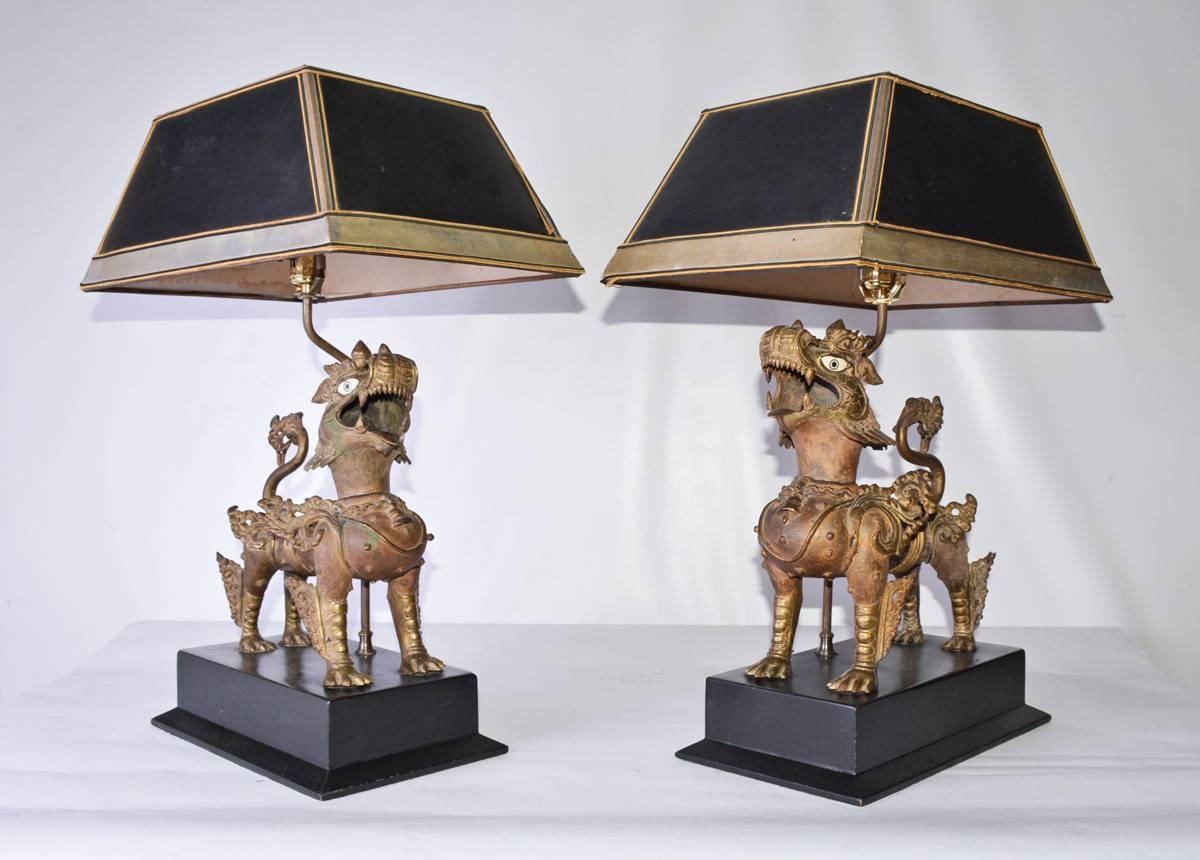 A bestial pair of Chinese brass Fu dogs with elaborate ornate details have been transformed into table lamp bases standing on black wood bases and sheltered by black and silver lamps shades.

Wired post & socket, 17
