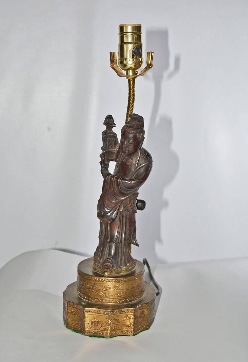 La lampe vintage a comme base une figure chinoise drapée en bois sculpté se tenant sur une base métallique gaufrée à deux niveaux. La lampe est électrifiée pour une seule ampoule américaine.
