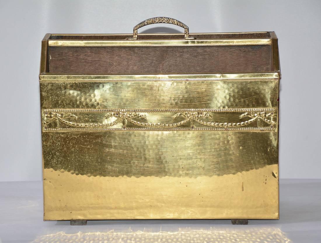 The vintage magazine basket is made of hammered brass sheeting on the front and sides nailed to a wood base with a brass handle top centre. A band of festooning and bows runs across the front. Wood runners are attached to the bottom.