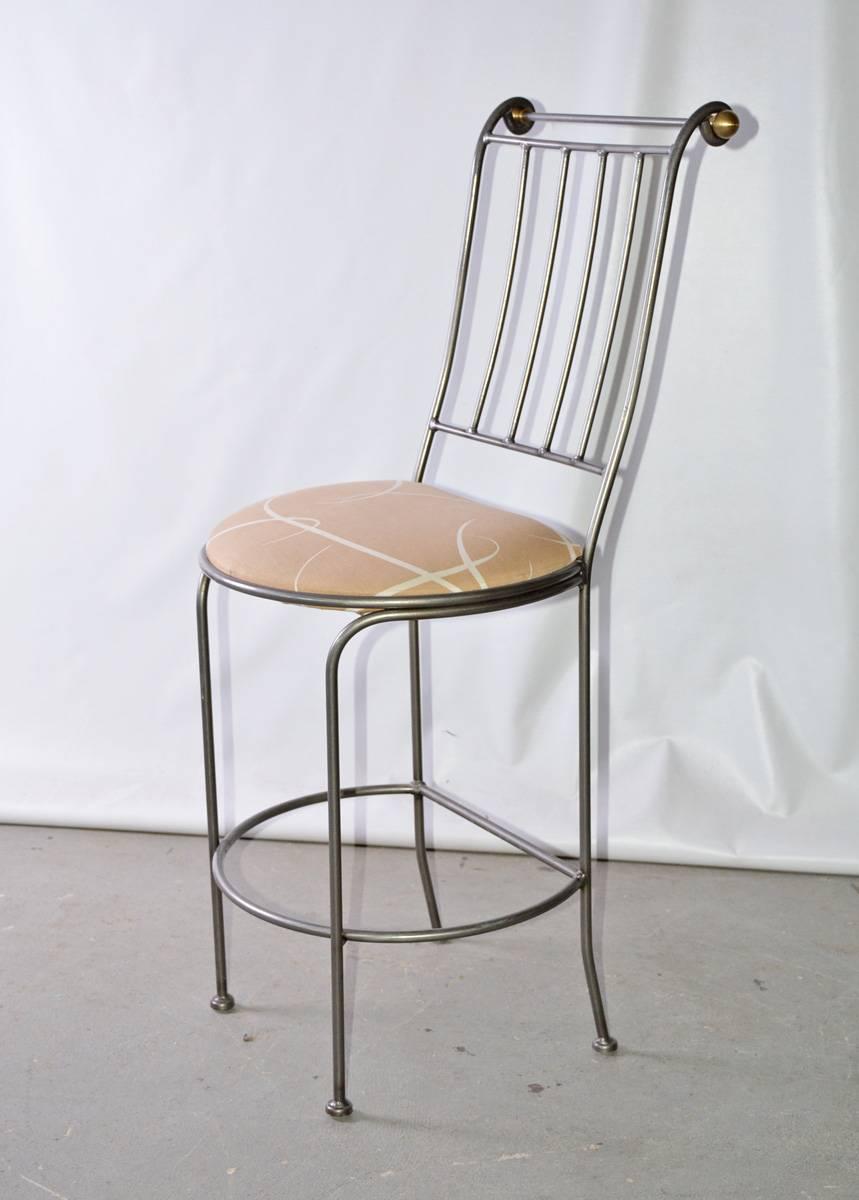 The contemporary bar stool is made of stainless steel bars and has round brass finials at the top of the back. The padded seat is upholstered in beige patterned cotton. 

 