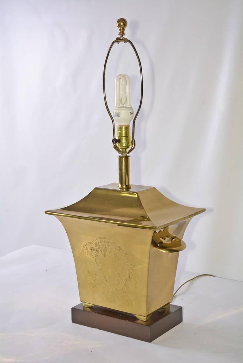 The brass and wood table lamp is in the Chinese pagoda style with engraved figures and animals in a pastoral setting and repeated on both sides. The brass pot with handles sits on a stained wood base. Wired for US use and a single bulb.