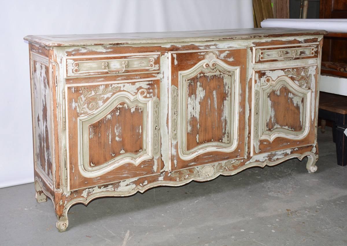 The antique server is in the French provincial, Louis XV style with cabriole legs and Rococo door panels. The doors have hand-carved flowers and leaves. The storage space includes two drawers and three cupboards with shelves. Doors have metal