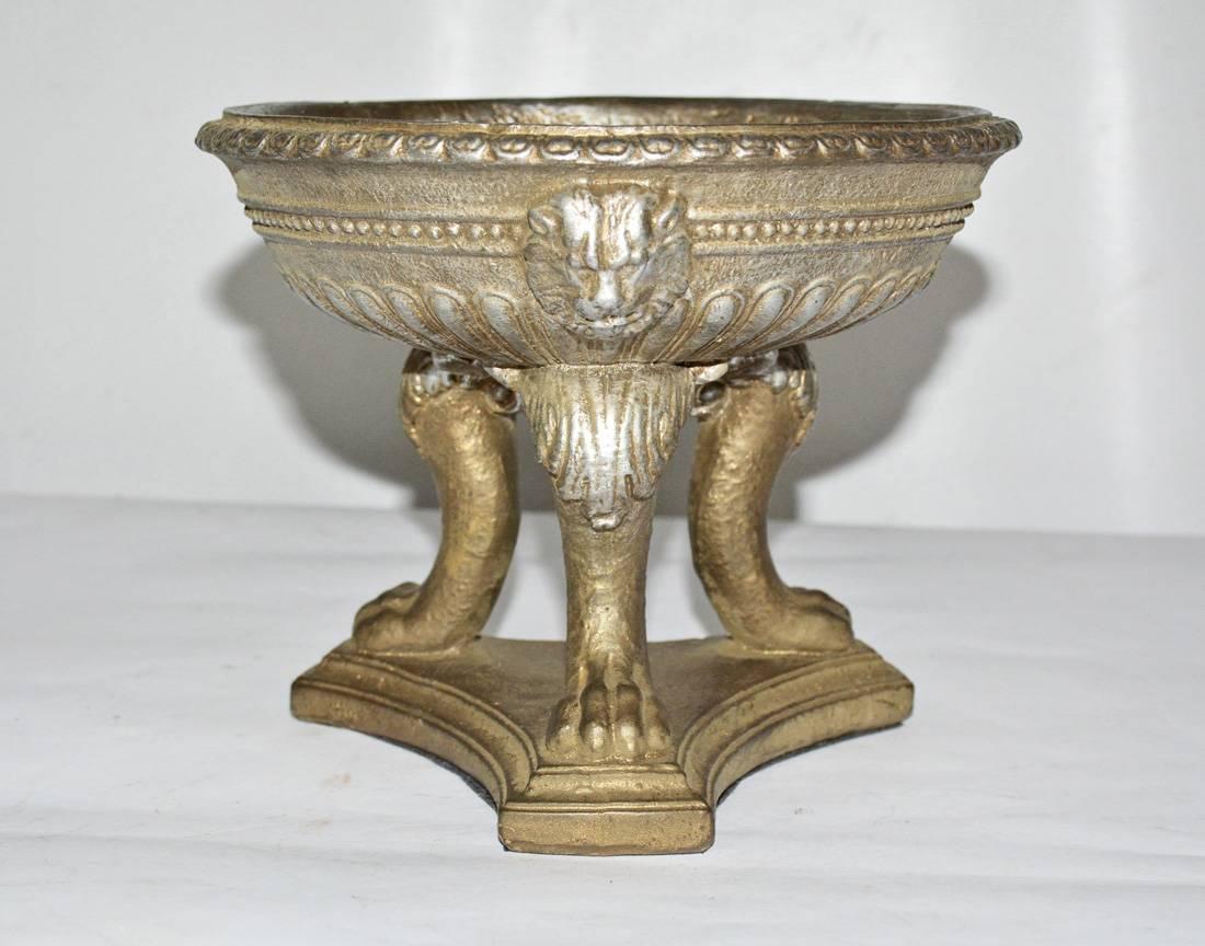 The vintage urn in the classical style sits on three animal legs attached to a triangular base. Three lion masks embellish the bowl. The urn is made of molded resin and is padded with felt. Label on the bottom reads Milano Designs. Wonderful as