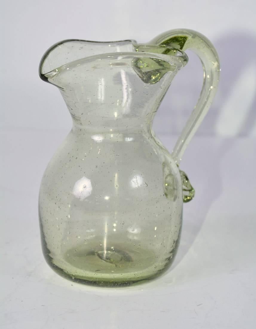 The vintage green glass creamer pitcher was handblown in Jamestown, VA, replicating the 17th century techniques in its production. The opening at the top is heart shaped. Other center circle on the bottom indicates the pontil mark where the piece