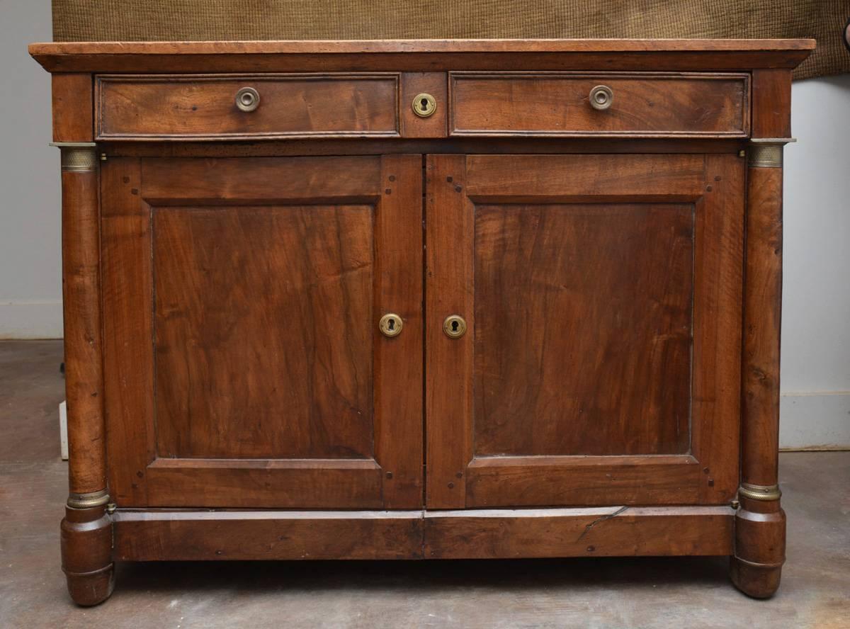 19th century or earlier Provincial Empire style walnut buffet sideboard or entry hallway cabinet has single top drawer with divided interior and base.  The capital of the two supporting side columns have cast brass decorations.  Interior has a
