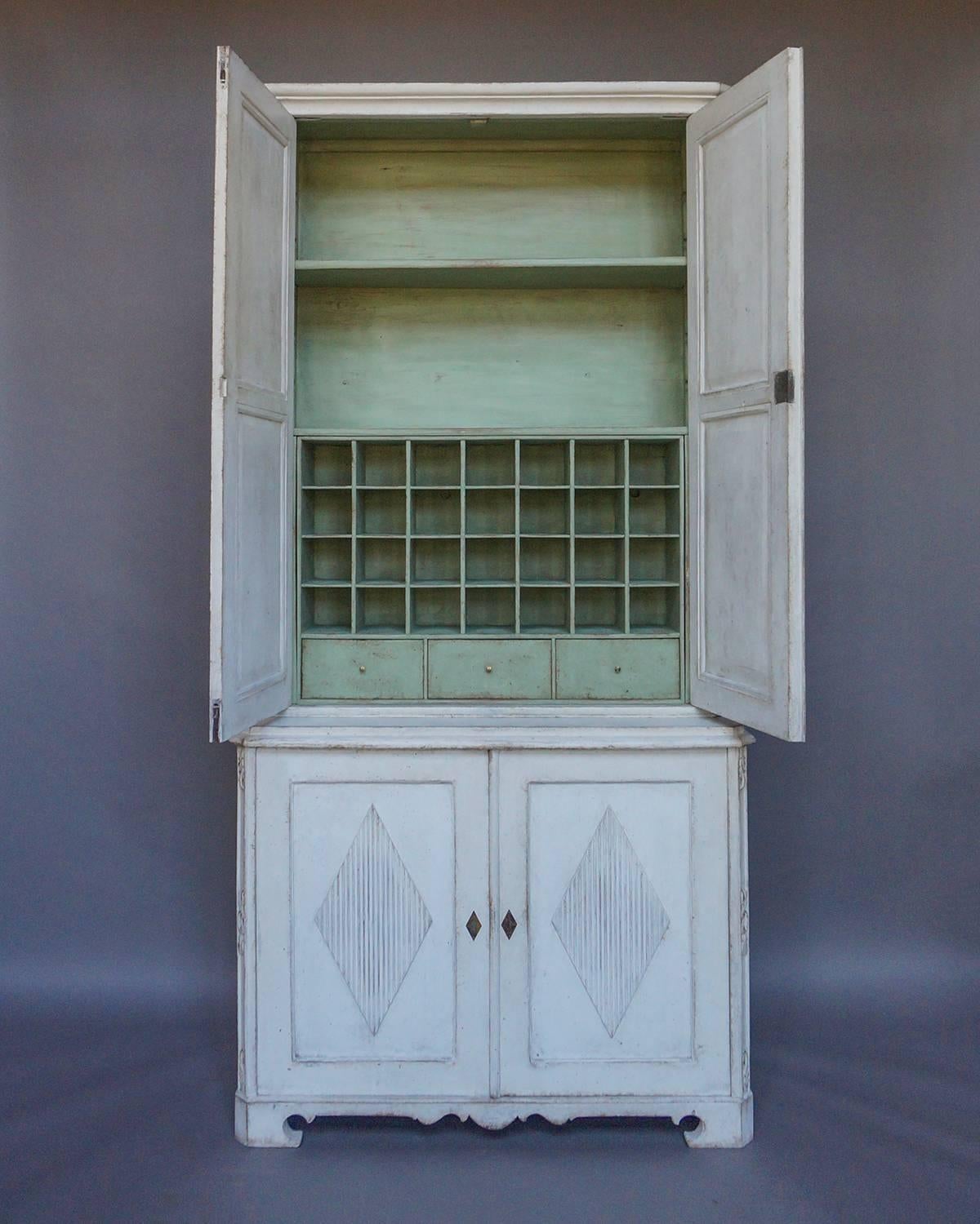 Cabinet in two parts, Sweden, circa 1800. The tall upper section has double doors, each with two reeded lozenges, which open onto two shelves, three small drawers, and 28 compartments. The deeper lower section has two shelves behind double doors,