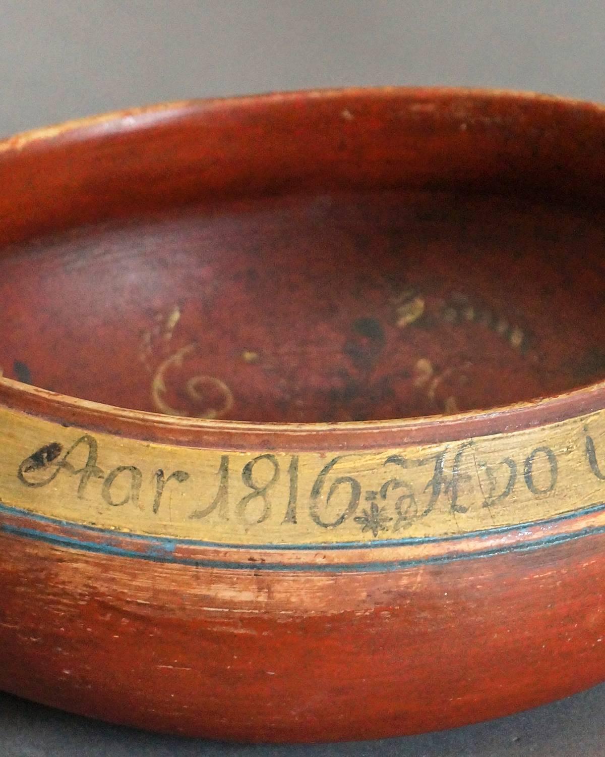 Turned wooden drinking bowl from Denmark painted a rich salmon and dated 1816. Around the rim is inscribed a verse translated as “He will not gather, but always drink! Wealth in the world he will not have. Year 1816.”  The interior is decorated with