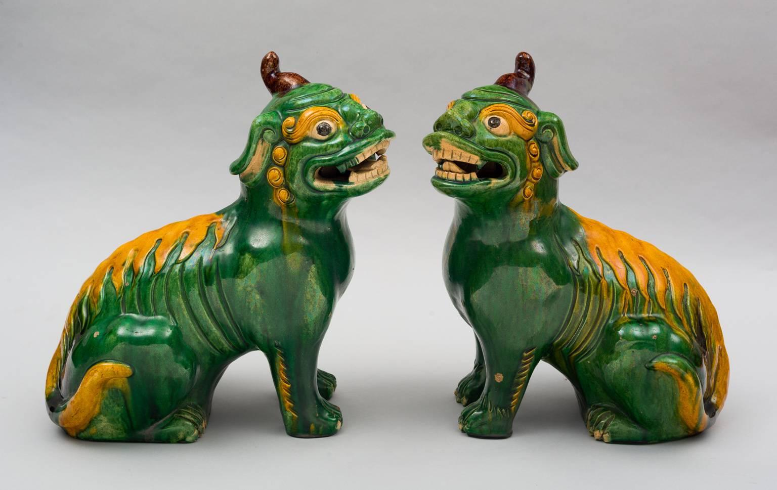 Pair of Chinese glazed pottery free standing, wild looking mythical beasts with open mouths, teeth, lolling tongues, bulging eyes and a single brown horn atop their heads. Polychrome decoration of a yellow fire pattern down their backs on a green