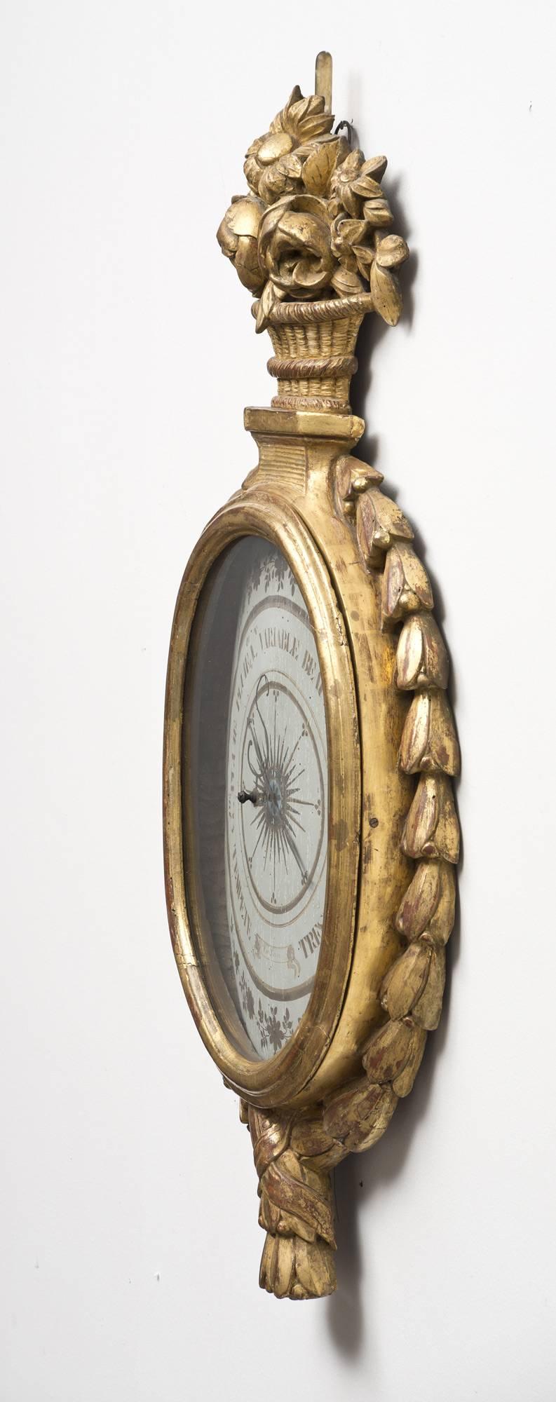 18th century French giltwood oval barometer by Torre, the crest a carved basket of flowers, the sides with cascading laurel leaves crossing at bottom and forming tassels. The grey painted and gilded dial with S-shaped hand is signed at bottom,