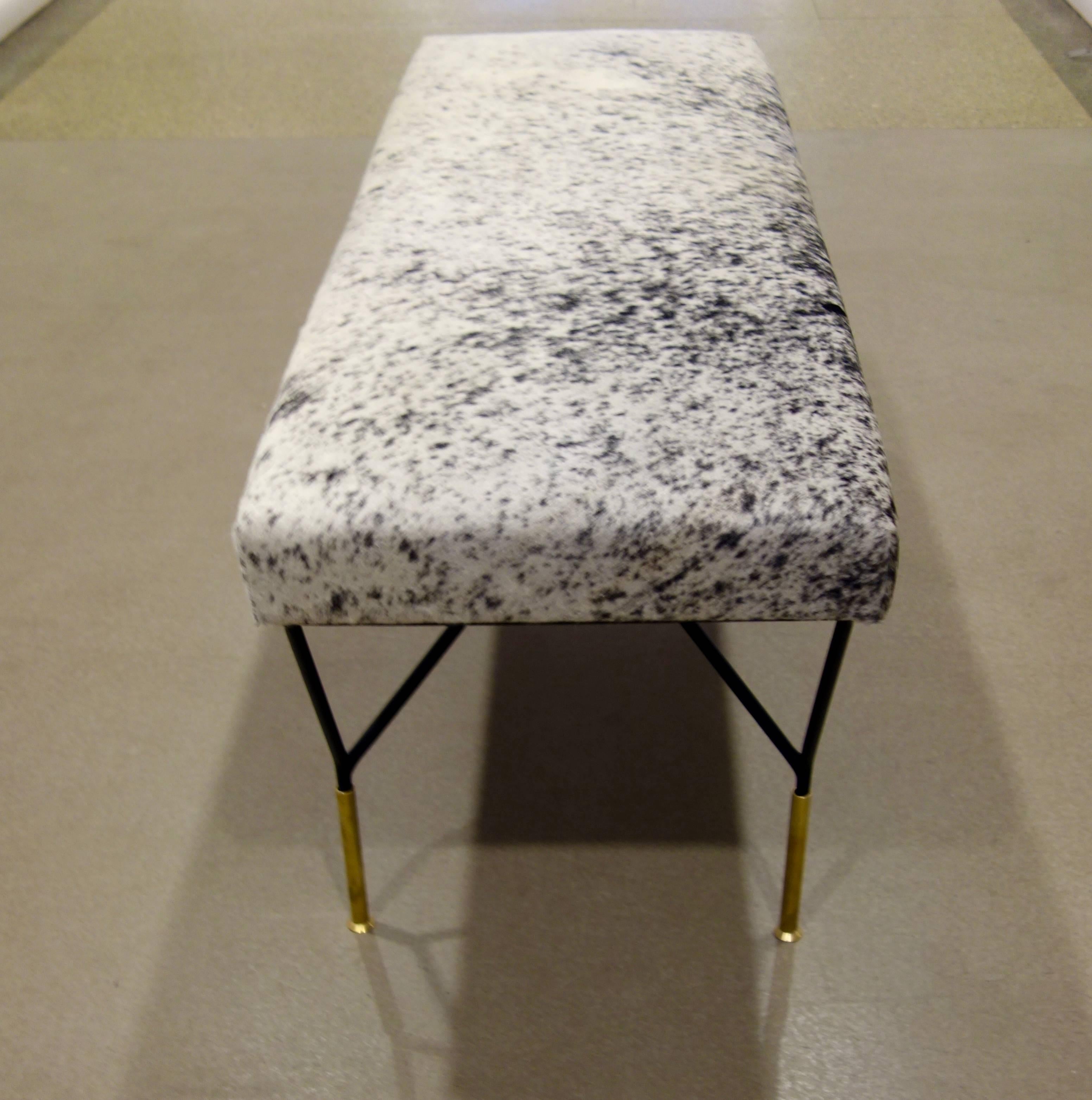 An Italian Mid-Century bench, the top newly upholstered in black and white cowhide, resting on black metal legs each attached at two points from the underside of the seat forming a triangle, ending in a single, elongated brass foot. 
Another bench