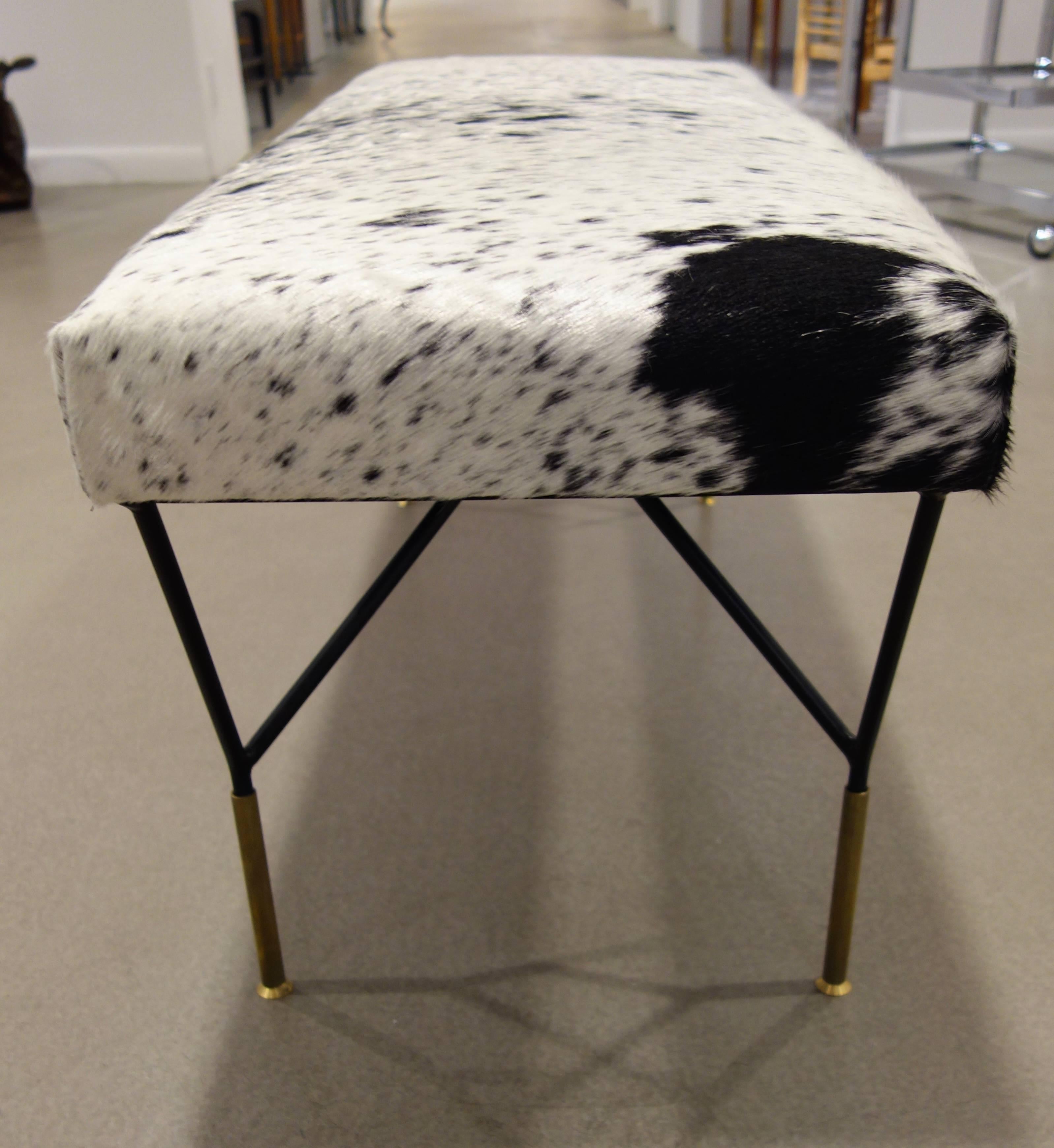 An Italian Mid-Century bench, the top newly upholstered in black and white cowhide, resting on black metal legs each attached at two points from the underside of the seat forming a triangle, ending in a single, elongated brass foot.
Another bench