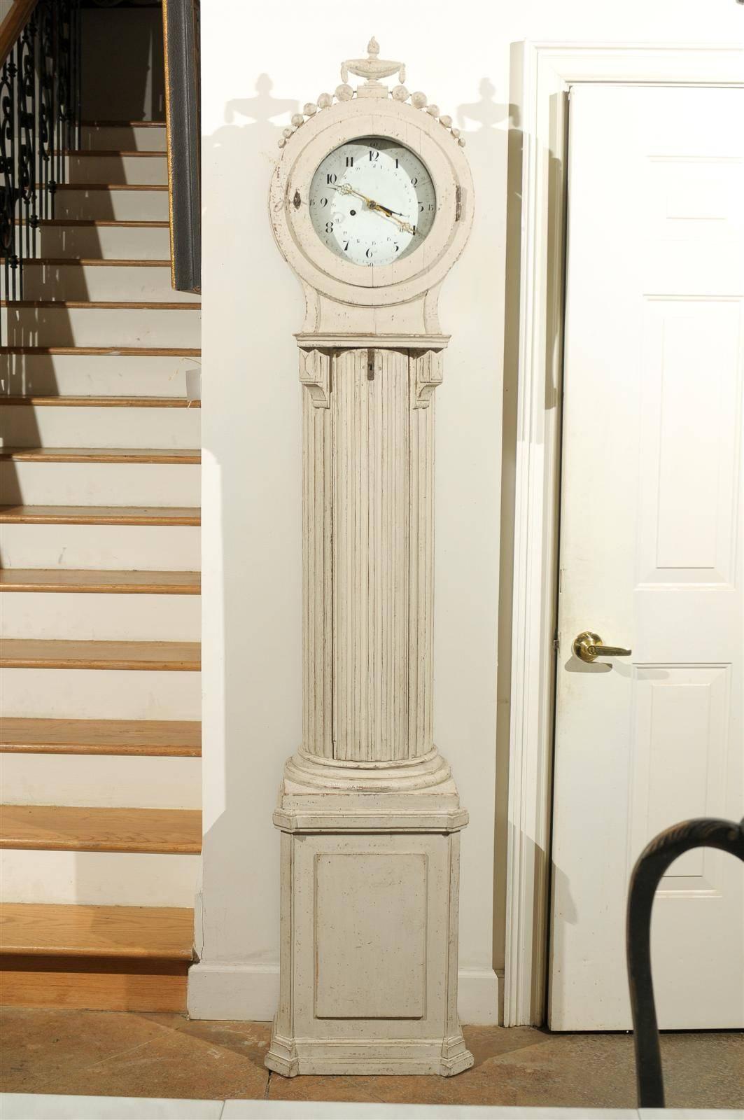 A Swedish painted tall case column clock from the mid 19th century, with carved crest, original movements and original paint. This exquisite Swedish clock features a delicately carved crest, adorned with a fire urn, accented with a swag and