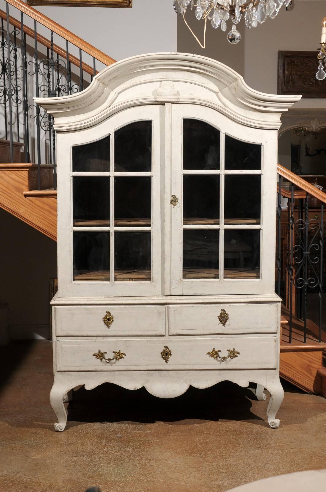 A Swedish period Rococo painted wood cabinet with bonnet-shaped pediment, glass doors, lower drawers and original hardware from the mid 18th century. Our eye is immediately attracted to the curvy molded bonnet top, beautifully crowing the confirming