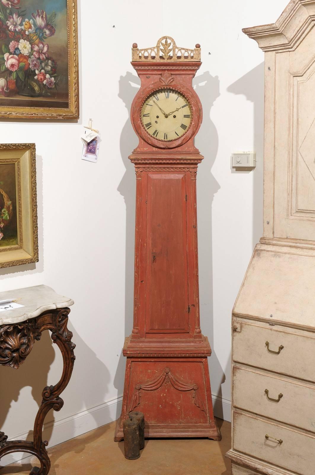 A Swedish Neoclassical tall case clock from the early 19th century with original orange paint and mechanisms. This Swedish longcase painted clock (commonly referred as a Mora clock) features an exquisite carved crest with pierced arched motifs