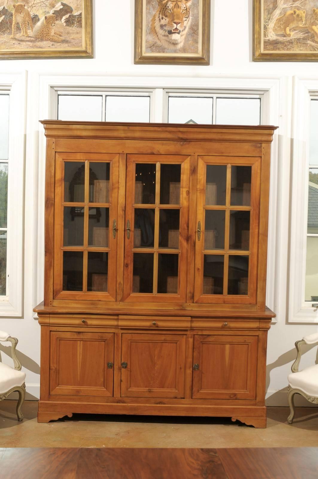 A French cherry wood two part bookcase with glass doors over three drawers and three doors from the early 20th century. This French cherry cabinet features a molded cornice sitting above three inset glass doors. Each door opens to reveal three inner