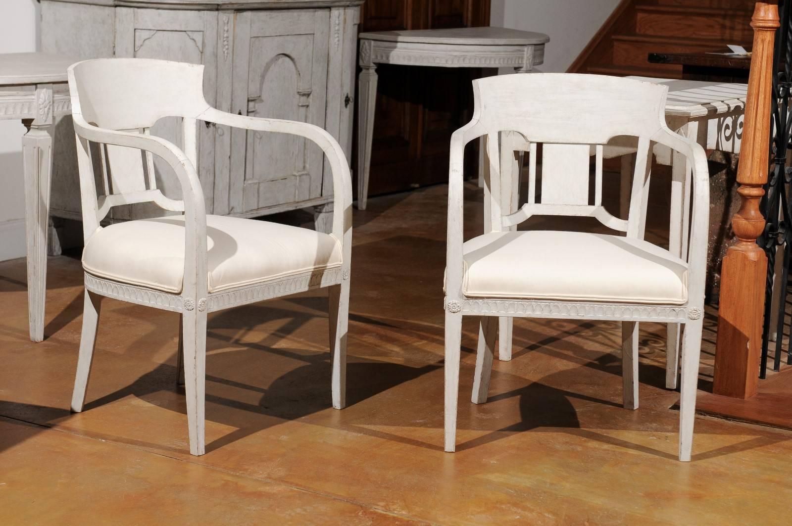 A pair of Swedish painted wood neoclassical style armchairs from the second half of the 19th century with tall open armrests, carved skirt and newly upholstered seat. Each of this pair of Swedish armchairs features a delicately curved back with