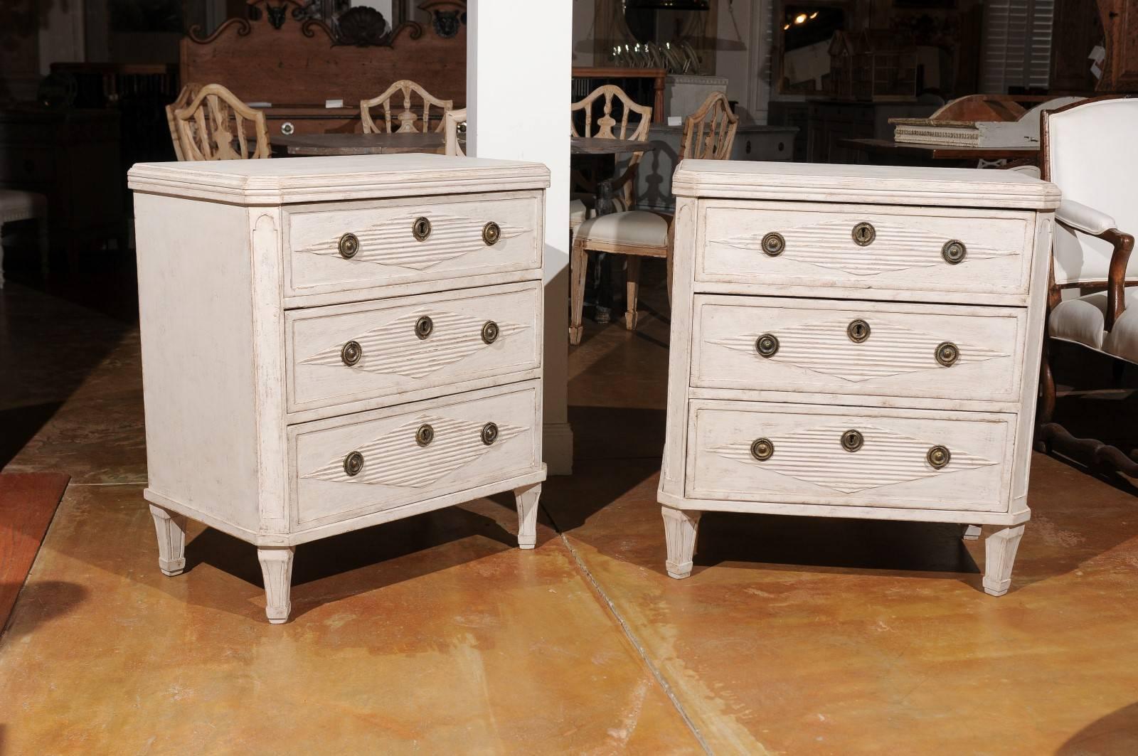 This pair of Swedish Gustavian three-drawer painted commodes from the mid-19th century features rectangular tops with canted corners in the front over three drawers each. The drawers are beautifully adorned with an elongated reeded diamond motif. A