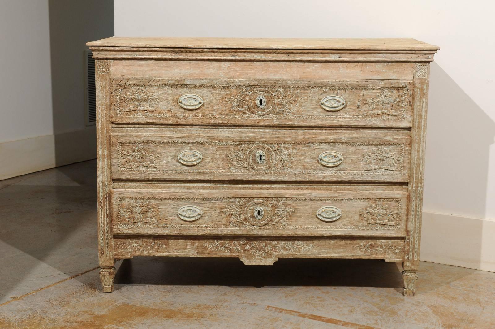 This exquisite French Louis XVI period bleached oak commode from the late 18th century features a three-plank rectangular top over three beautifully carved drawers. Each drawer is adorned with delicate floral motifs flanking the central escutcheon