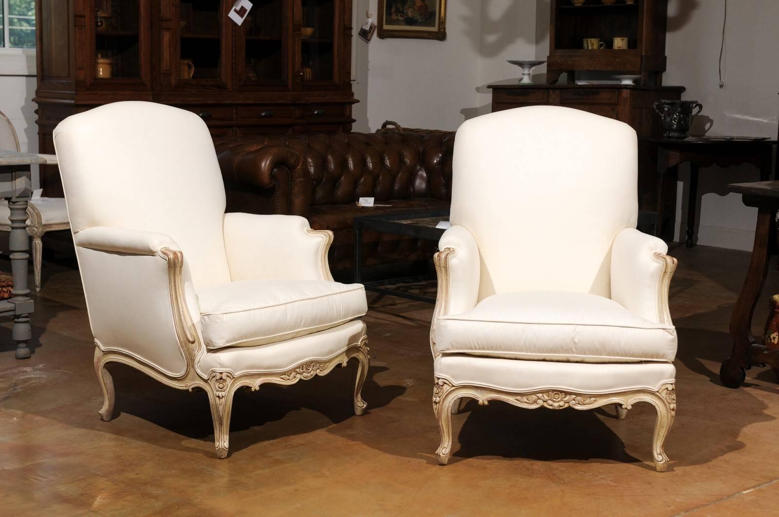 A pair of French Louis XV style painted wood bergère chairs from the early 20th century with carved skirt and new upholstery. Each of this pair of French painted bergères features a slanted back, flanked by two fully upholstered arms with scrolled