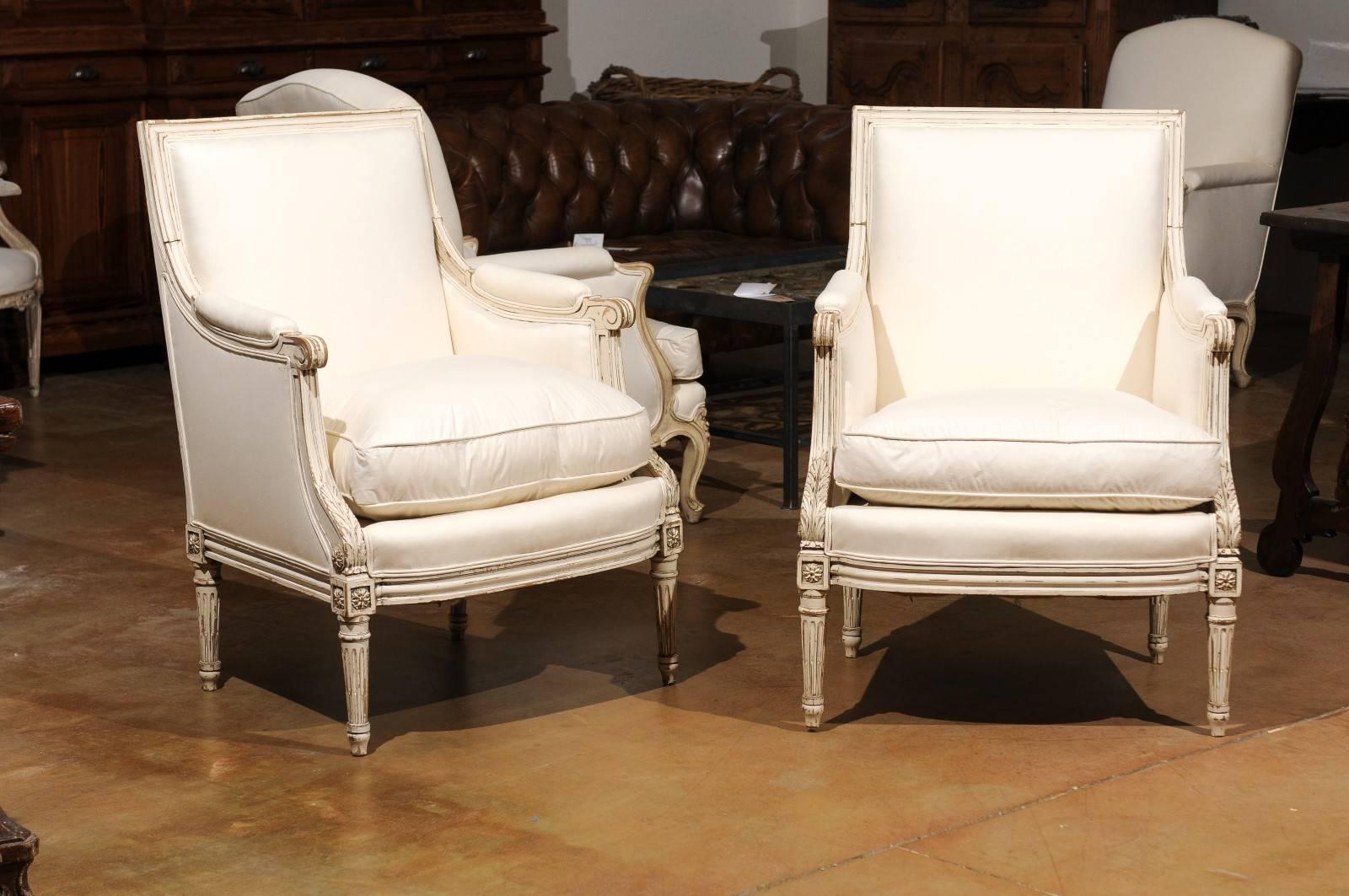 A pair of French Louis XVI style painted and carved wooden bergère armchairs from the early 20th century, with scrolled arms, fluted legs and new upholstery. Each of this pair of French Louis XVI style bergères features a rectangular, slightly