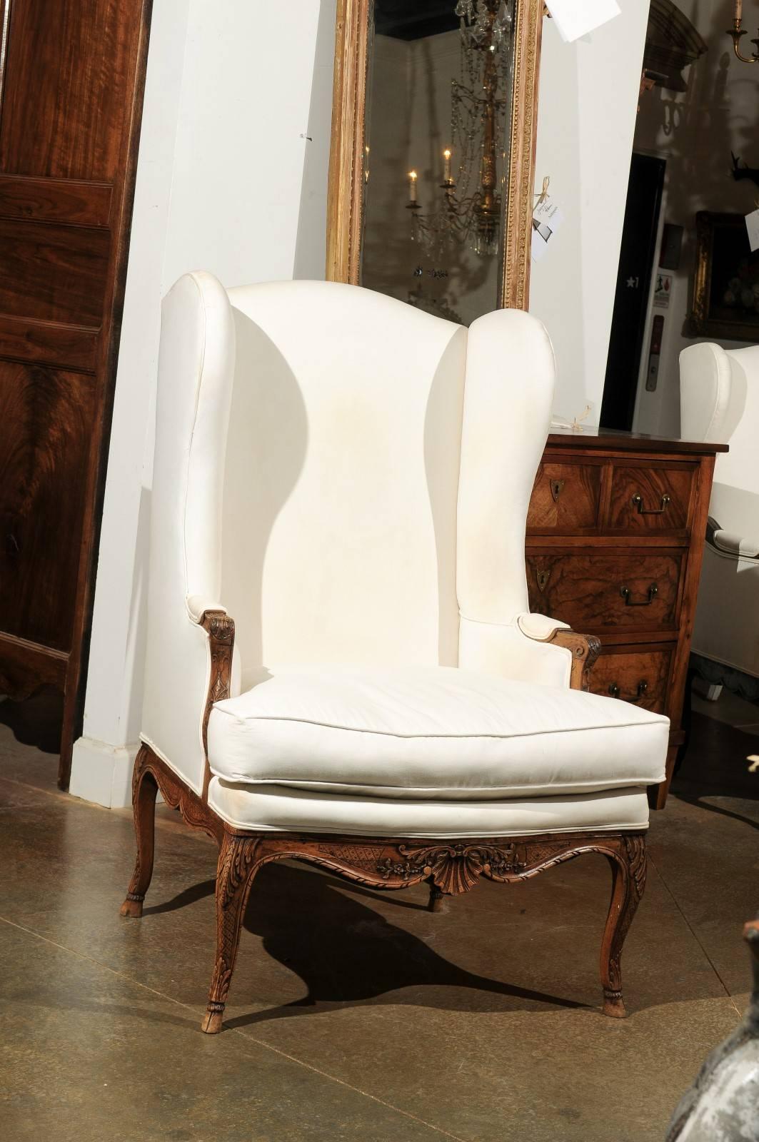 This French Louis XV style wingback chair from the early 19th century features a sturdy cotton upholstery with double welting over a delicate carved walnut body. The wingback, intended to provide the sitter with a sense of privacy, evolves into