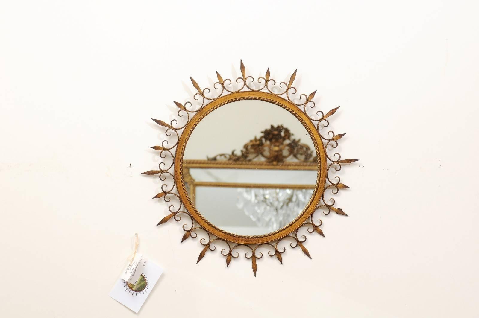 A French forged iron gilded sunburst mirror with stylized fleur-de-lys and braided molding from the midcentury. This French sunburst mirror was born in the 1950s and features a clear mirrored glass in the center, delicately surrounded by a braided