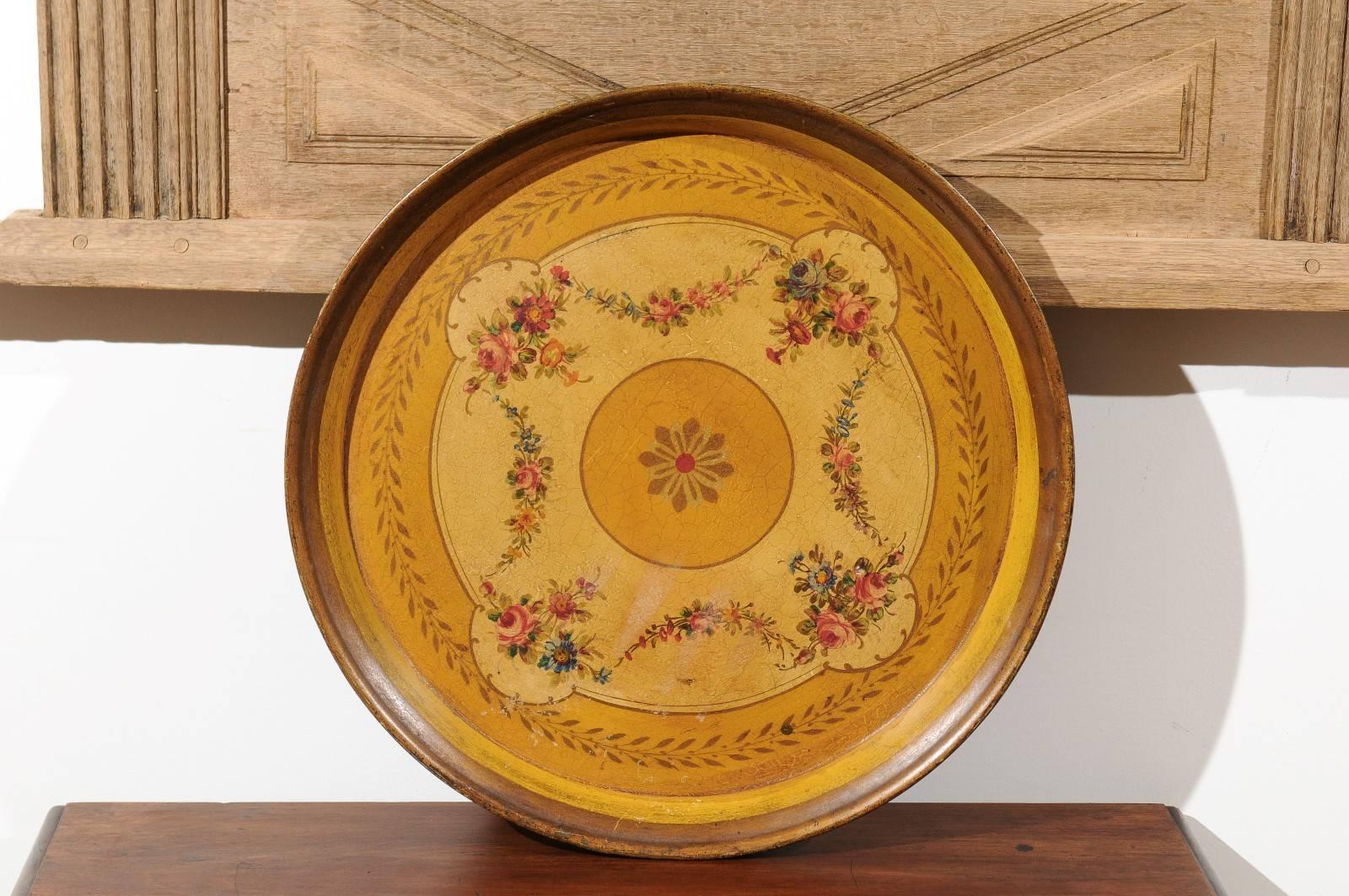 A French round painted tole tray with floral motifs from the 19th century. This French circular tole tray is adorned on the surround with a delicate foliage motif reminiscent of olive branches. The center is decorated with a stylized floral motif