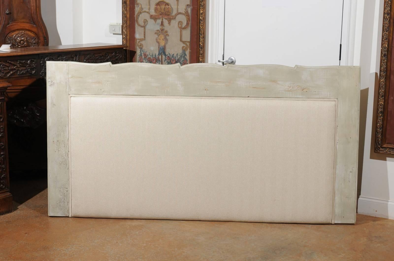 A painted wood upholstered headboard perfect for a king-size bed, made from the shelf of an antique French bookcase. This headboard features a light grey / green painted wooden frame, topped with a delicately carved upper rail with serpentine lines.