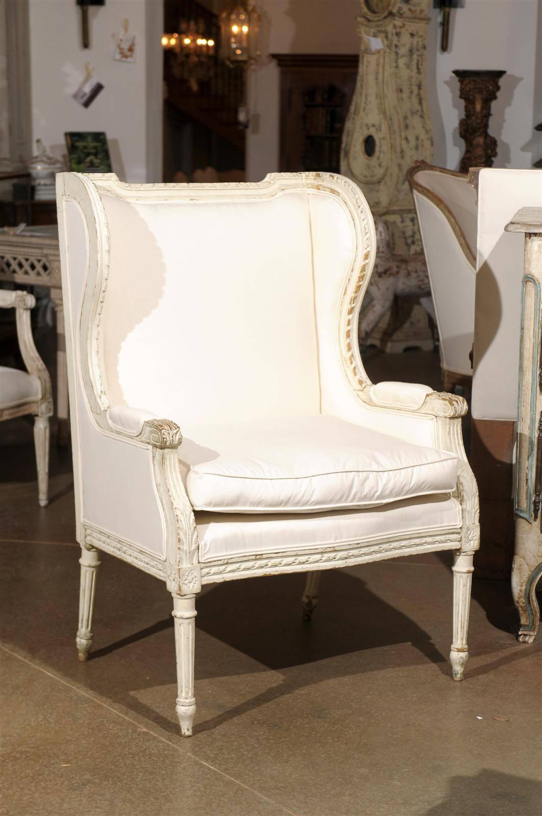A French Louis XVI style painted wood upholstered bergère chair from the early 20th century. This French bergère à oreilles (wingback chair) features a straight back with curved sides, padded armrests and scrolled knuckles. The frame is adorned with