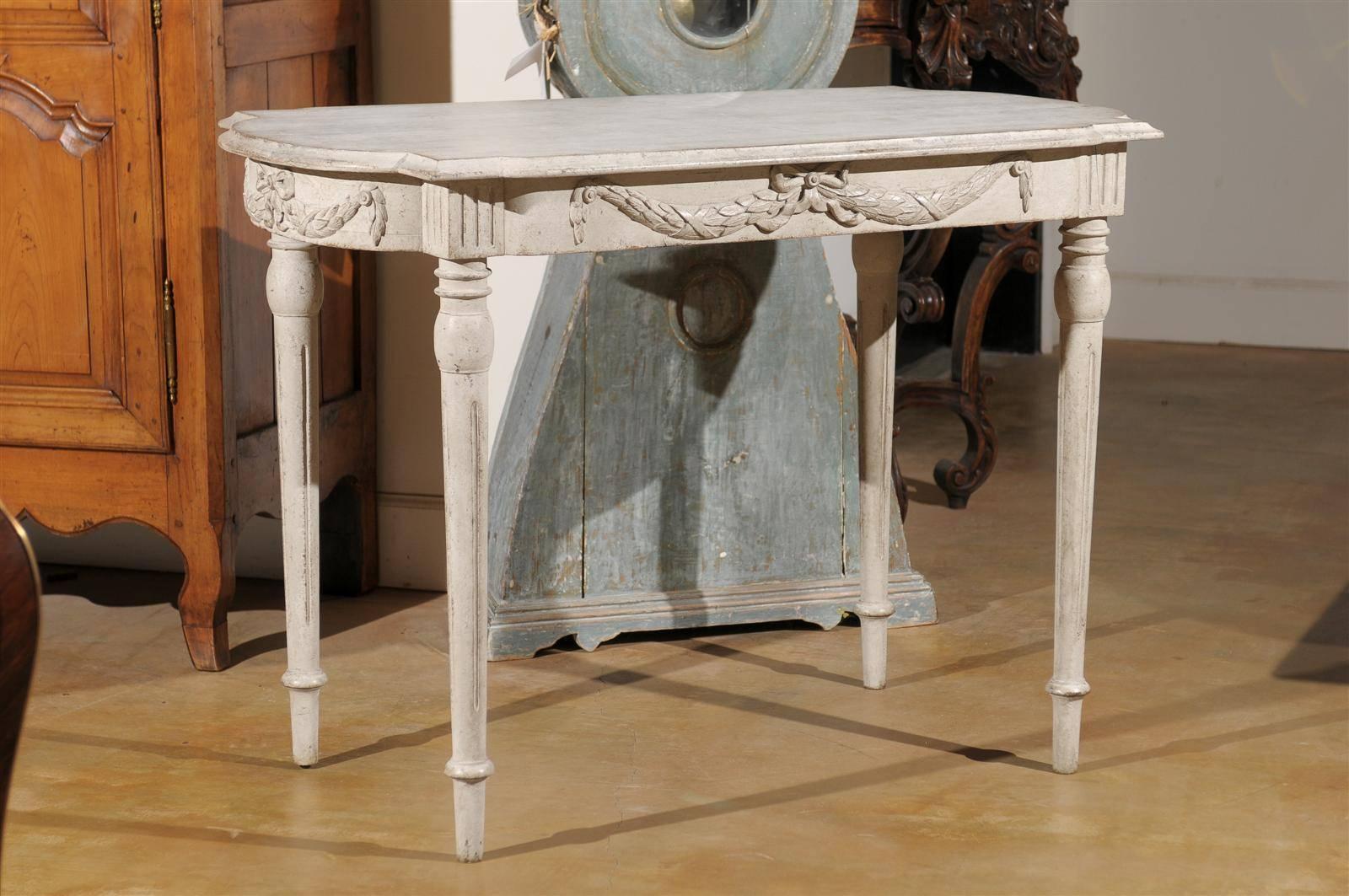 This Swedish painted wood Gustavian Style tea table from the late 19th century features a contoured and beveled top over an exquisitely carved apron. Each side is adorned with a delicate swag and ribbon. The fluted knees are raised on four elegant