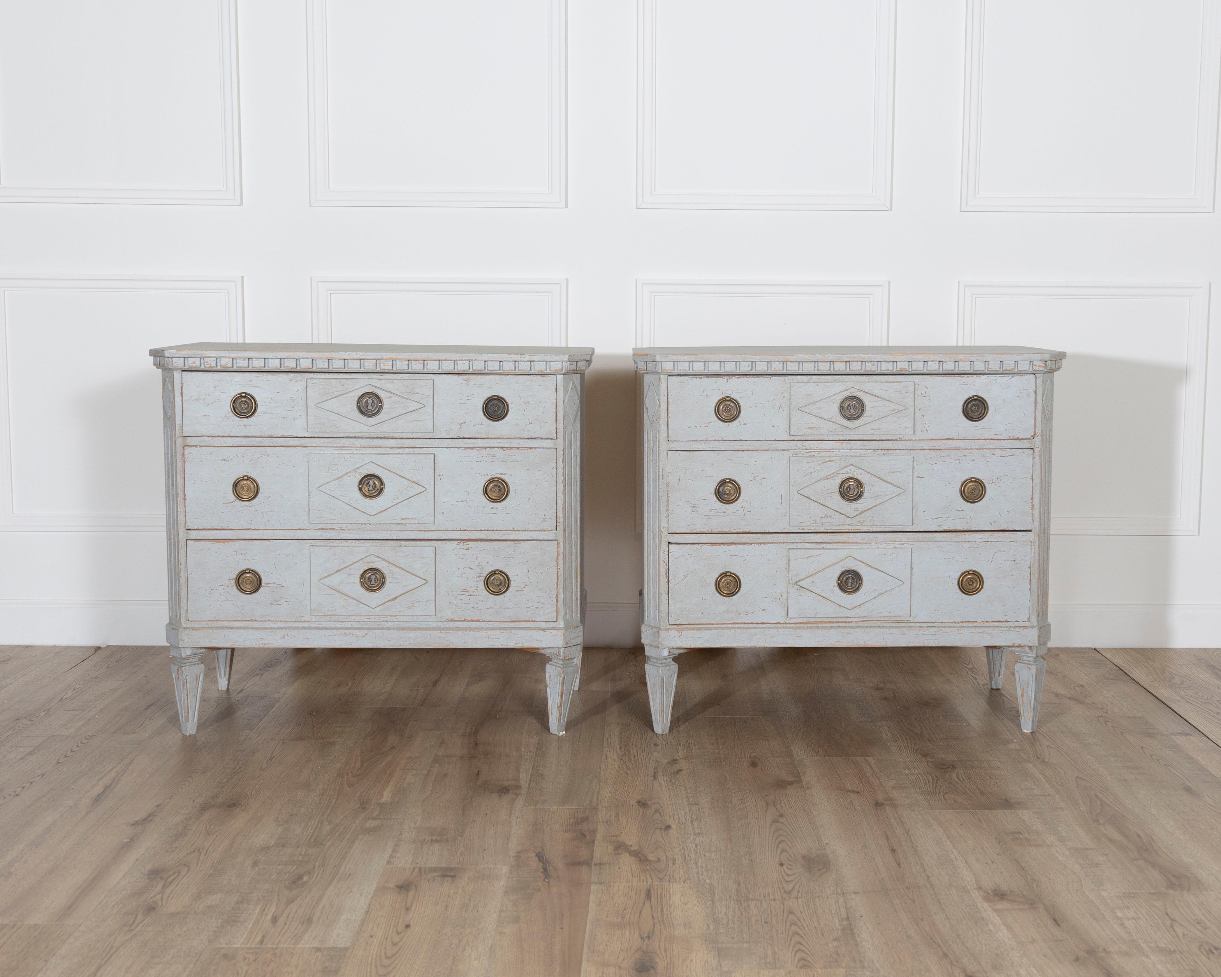 A graceful pair of Swedish Gustavian-style chests of drawers, crafted circa 1920 and finished in a pale, softly distressed grey-white paint with warm undertones. Each chest features three spacious drawers adorned with carved diamond-shaped paneling