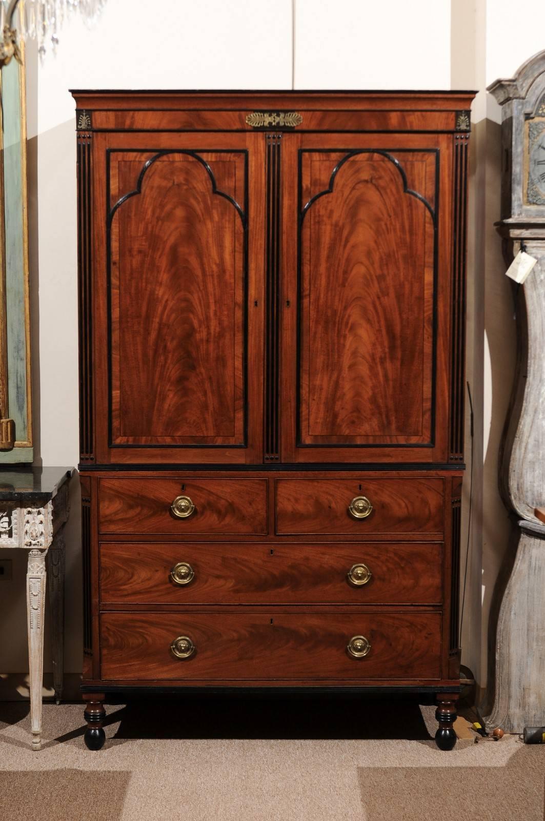 Regency gilt-metal mounted and part ebonized mahogany linen press with arched paneled doors enclosing three sliding shelves, four drawers below and turned feet.