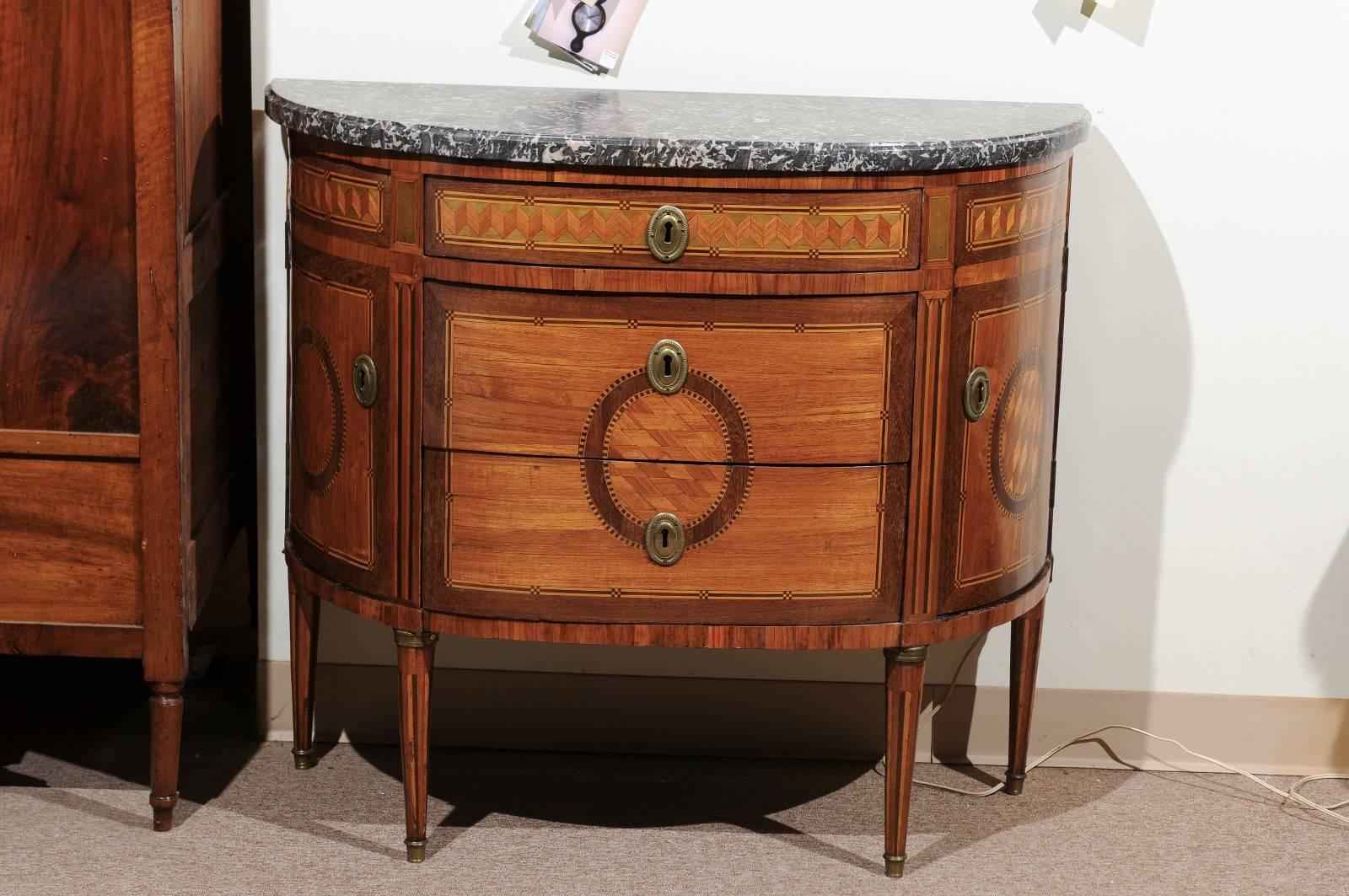 Louis XVI period parquetry inlaid commode in demilune shape with three drawers flanked by two cabinet doors with fitted interior and grey marble top, France, 18th century. The inlay in boxwood and ebony on the legs and beside the drawers creates the