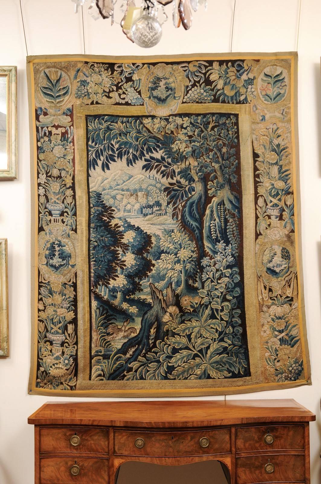 A 18th century French aubusson tapestry featuring foliage boarder with center oriented landscape scene in teal blue, brown and tan hues.