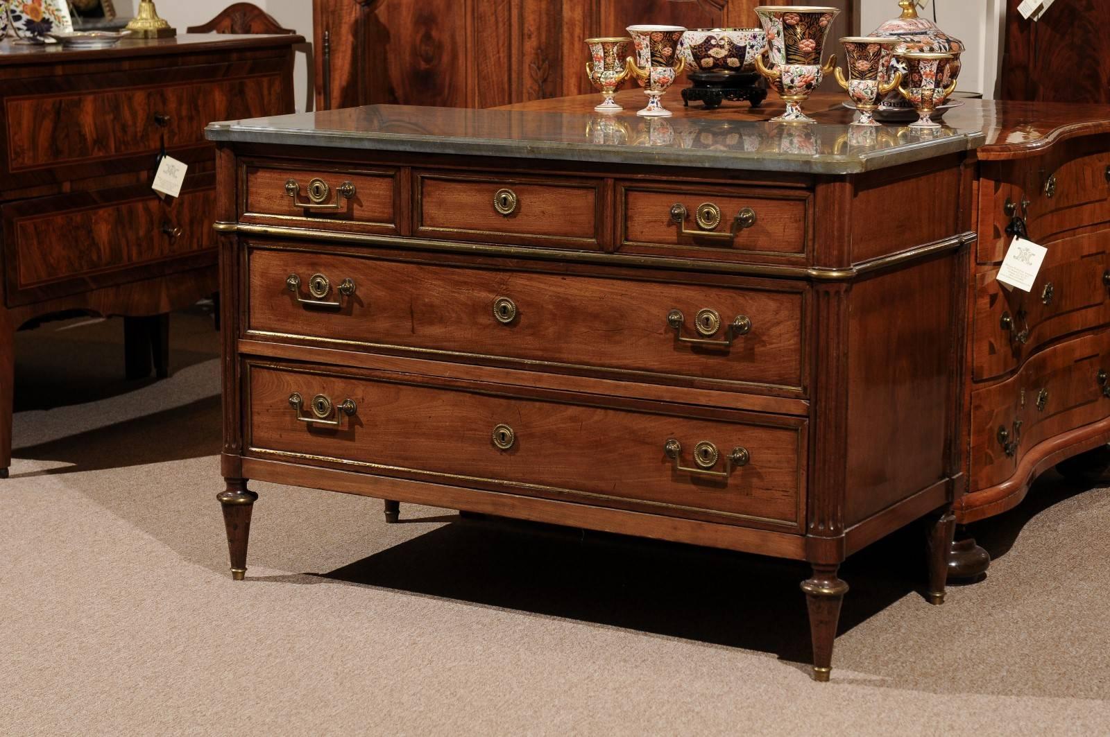 The Louis XVI commode in a medium walnut with grey marble top, rounded fluted corners flanking 3 short drawers and 2 long drawers below with brass pulls and brass inlay. All supported by turned tapered legs. 





