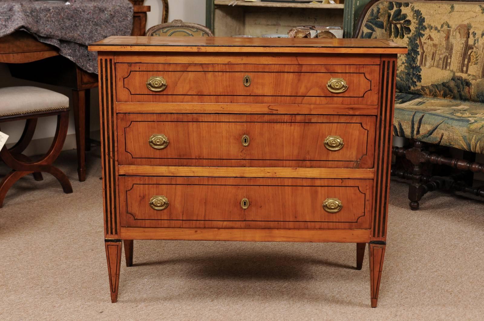 The neoclassical Italian fruitwood commode of shallow size with ebonized inlay, three drawers with brass pulls and tapered legs.