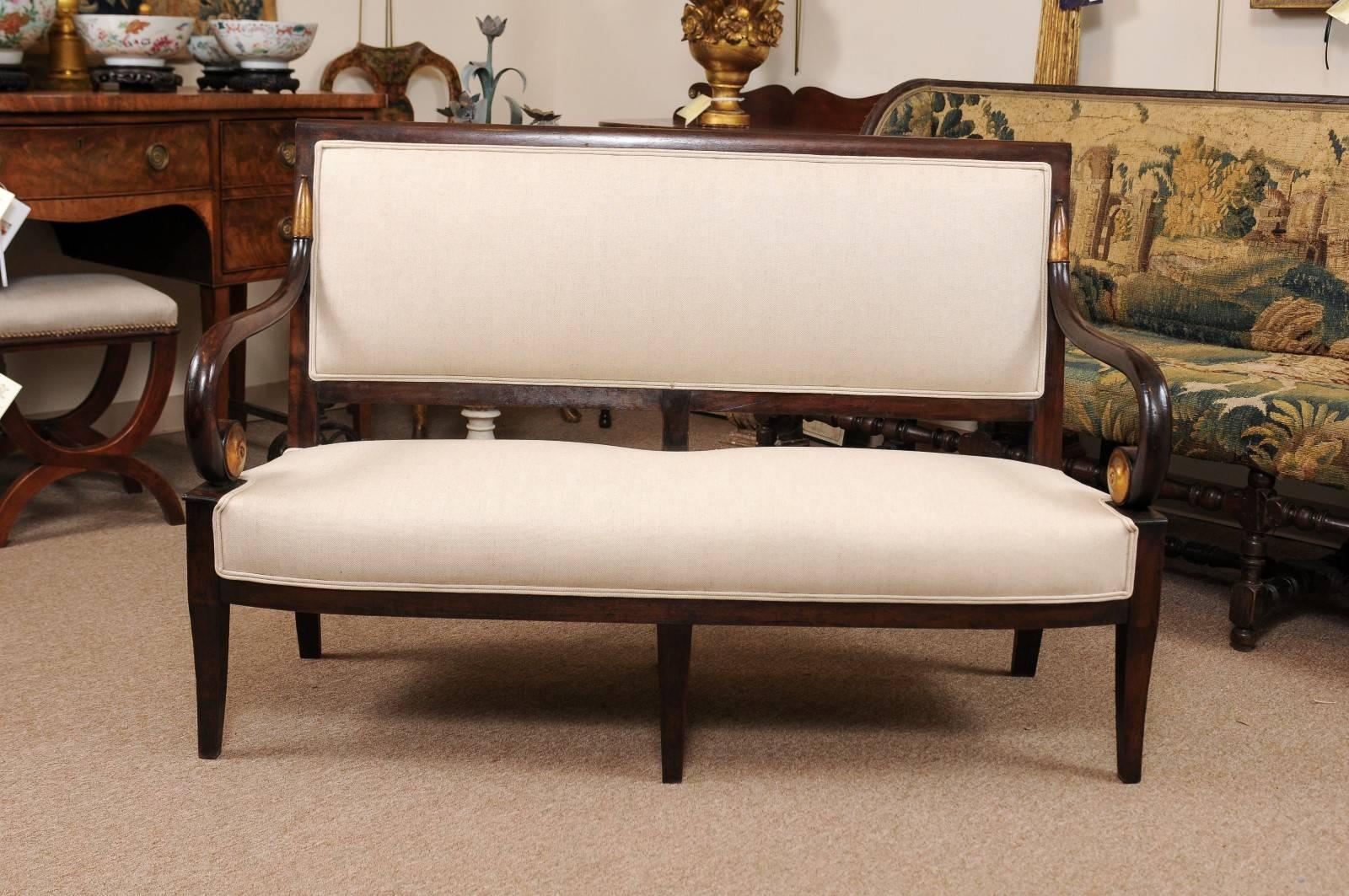 Empire Period Settee in Mahogany with Scroll Arms featuring Gilt Accents and Splay Legs, France circa 1820. The wood finish is in a dark chocolate hue juxtaposed against the light beige linen upholstery (new).