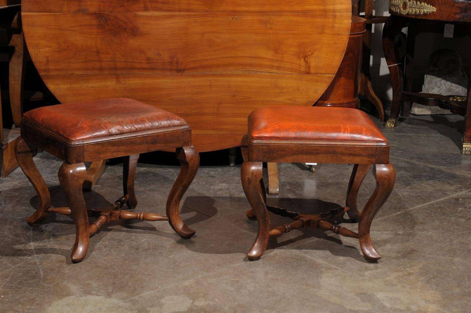 A pair of Italian late 19th century wooden stools with old leather seats.  This pair of wooden Italian stools from the late 19th century features a square top covered with leather, and four curved legs linked together by two trestles forming a